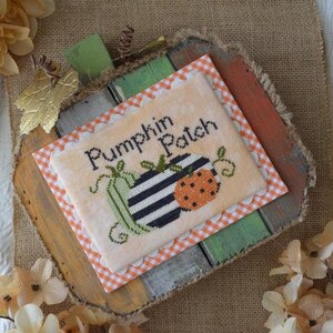 May include: A handmade, rustic wooden pumpkin-shaped sign with a cross-stitch design that reads "Pumpkin Patch." The cross-stitch features a striped pumpkin and a dotted pumpkin in orange and black. The sign has a gingham border.