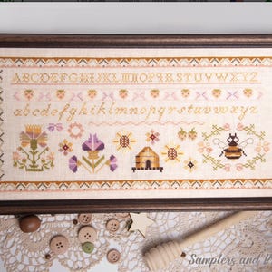 May include: A rectangular framed cross-stitch sampler on light fabric featuring an alphabet in uppercase and lowercase letters, rows of strawberries, flowers, a beehive, and a bee. The border has a geometric pattern in brown and orange.