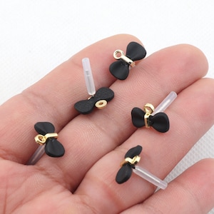 May include: Five black bow-shaped charms with gold accents and clear plastic posts. The charms are on a hand.