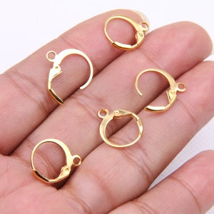 May include: Five gold-toned earring hooks, also known as earring findings, are displayed on a hand. The hooks are made of metal and have a smooth finish.
