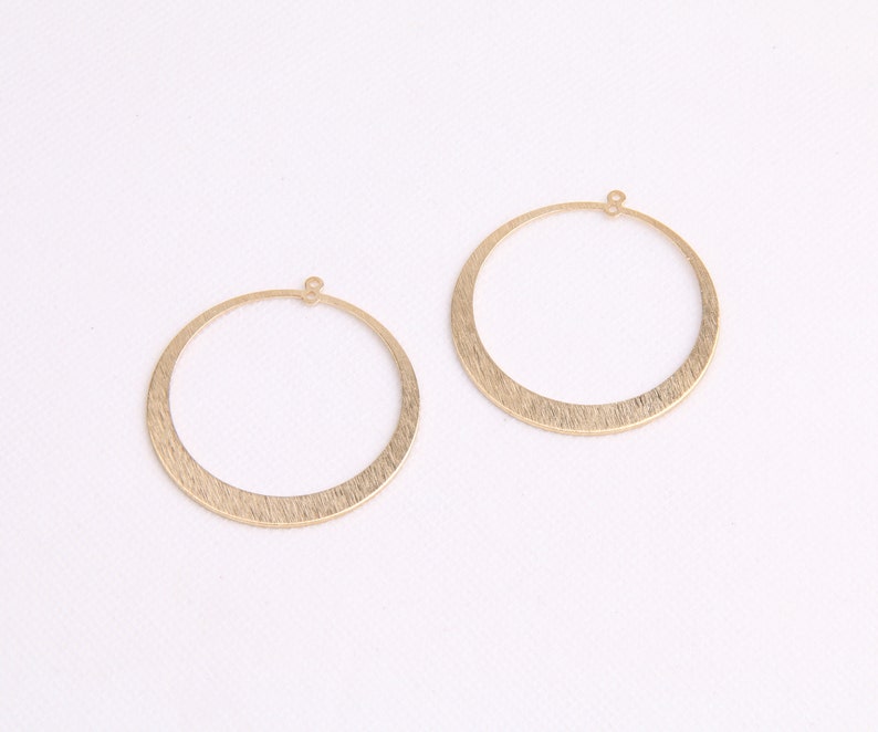 May include: Two gold-toned hoop earrings with a brushed finish. The hoops are circular and have a small loop at the top for attaching to an earring post.