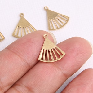 Könnte beinhalten: Vier goldfarbene Metall-Anhänger in Fächerform mit einer Öse oben. Die Anhänger liegen auf einer weißen Oberfläche und eine Hand hält einen der Anhänger.