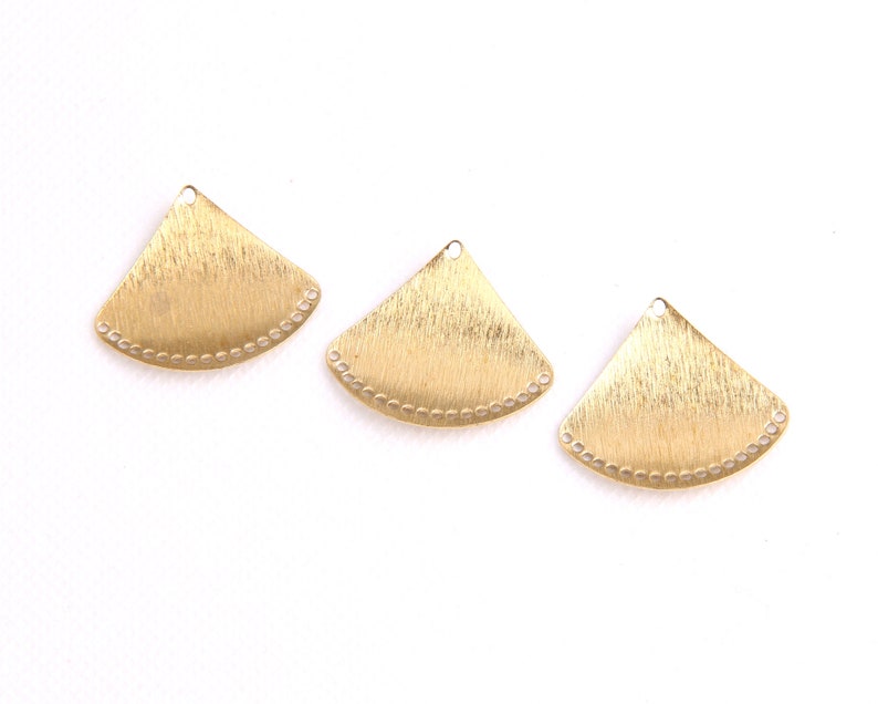 May include: Three gold-colored metal fan-shaped charms with a beaded edge. The charms have a brushed finish and a hole at the top for attaching to jewelry.