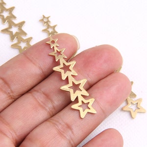 May include: Gold-colored star charms in various sizes, arranged in a cascading pattern. These charms are cut-out, with open star shapes. The charms are held in a hand against a white background. These charms are ideal for jewelry making.