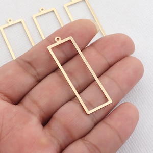 May include: Four gold-colored metal rectangular frames with a loop at the top. The frames are empty and appear to be for jewelry making.
