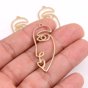 May include: Gold-colored metal face pendants with a minimalist design. The pendants feature a single line drawing of a face with an eye, nose, and a heart-shaped mouth. The pendants have a small hole at the top for stringing.