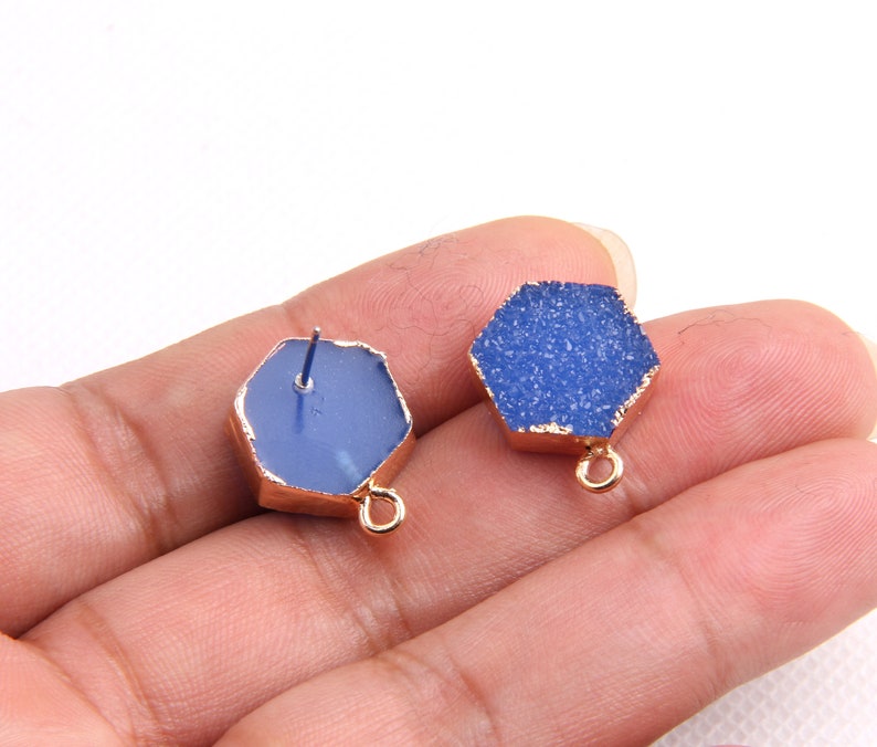 May include: Two blue hexagon-shaped gemstone pendants with gold-toned edges and a loop for hanging. The pendants have a textured, sparkly surface.