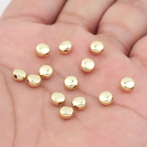 May include: A collection of 12 gold-colored round beads with a hole in the center. The beads are scattered on a hand.
