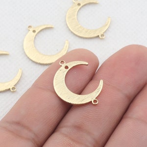 Puede incluir: Cuatro abalorios de metal de color dorado en forma de media luna con anillas de salto. Los abalorios están sobre una superficie blanca y una mano sostiene uno de los abalorios.