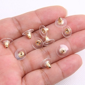 May include: A close-up of a hand holding a group of clear plastic earring backs with gold-colored metal posts. The backs are designed to be used with stud earrings.