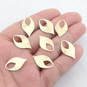 May include: A collection of eight gold-colored leaf-shaped charms. Each charm has a hammered texture and a teardrop-shaped cutout. The charms are arranged on a light background, suggesting they are jewelry components or craft supplies.