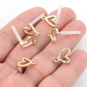 May include: Gold heart-shaped charms with a textured surface and a small loop at the top. The charms are attached to clear plastic posts.