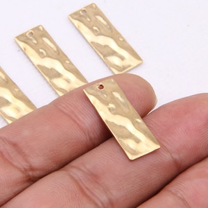 May include: Four gold-colored metal rectangular tags with a hammered texture and a hole at the top. The tags are on a hand.