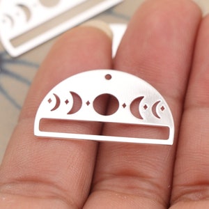May include: A silver-colored, half-circle pendant with a celestial design. The pendant features cut-out crescent moons, diamonds, and a full moon. A rectangular bar is at the bottom, and a small hole is at the top for stringing.