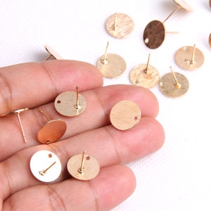 May include: A close-up of a hand holding a variety of gold-coloured earring blanks. The blanks are round and have a small hole in the centre. They are perfect for making your own earrings.