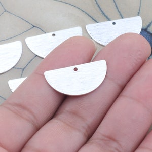 Puede incluir: Primer plano de una mano sosteniendo cinco amuletos de metal de media luna plateados con un agujero en la parte superior. Los amuletos son lisos y tienen un acabado cepillado.