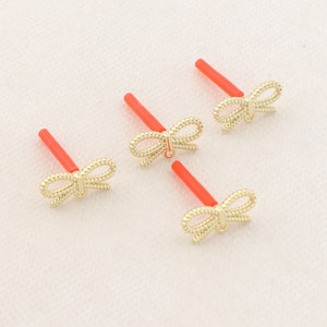 May include: Four gold bow-shaped nose rings with red plastic posts. The bows are made of a twisted wire design.