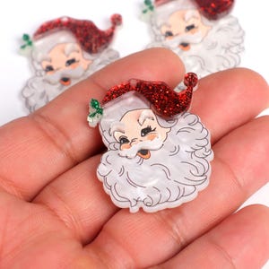 May include: Three white and red glitter Santa Claus resin ornaments with white beards and red hats. The ornaments are on a hand.