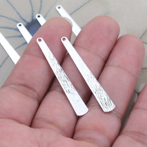 以下が含まれることがあります： テクスチャ加工が施されたシルバーメタルペンダントチャーム。チャームは長く、細長く、丸みを帯びたトップと、ジュエリーに取り付けるためのトップに穴が開いています。