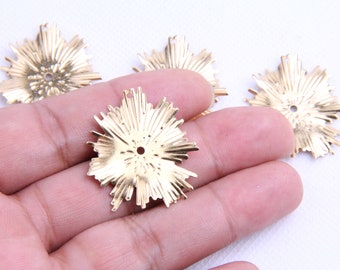 Puede incluir: Primer plano de varios dijes de metal dorados con forma de ráfaga de estrellas. Cada dije tiene un agujero central y líneas radiales, lo que sugiere un diseño de sol. Estos dijes probablemente se utilizan para hacer joyas u otros proyectos de artesanía.