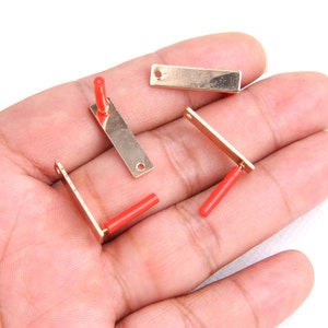 May include: Four gold-colored metal pieces with red plastic tips. The pieces are rectangular and have a hole at one end. They are lying on a hand.