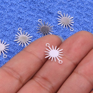 Może przedstawiać: Pięć srebrnych zawieszek w kształcie gwiazdy z otworem na górze do przymocowania do biżuterii lub innych prac ręcznych. Zawieszki znajdują się na niebieskiej, puszystej powierzchni, a jedna z nich jest trzymana w dłoni.