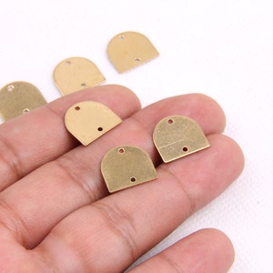 May include: Several small, brass-colored metal charms in a half-circle shape with two holes at the bottom and one at the top. The charms are scattered on a white surface, with some held in a hand.