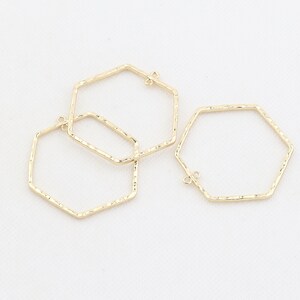 Op de afbeelding: Drie gouden, zeshoekige metalen hoepels met een getextureerd oppervlak. De hoepels hebben een kleine lus aan de bovenkant om aan sieraden te bevestigen.