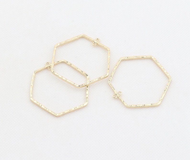 Op de afbeelding: Drie gouden, zeshoekige metalen hoepels met een getextureerd oppervlak. De hoepels hebben een kleine lus aan de bovenkant om aan sieraden te bevestigen.