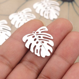 May include: White metal charms shaped like monstera leaves. The leaves have a cut-out design and are perfect for jewelry making or crafting.
