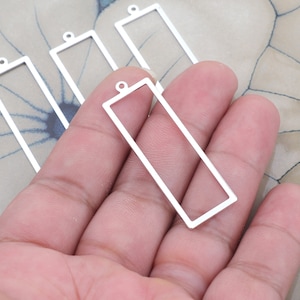 May include: Four silver-toned metal rectangular frames with a loop at the top. The frames are empty and appear to be for jewelry making.