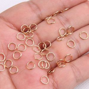 May include: A close-up image of a hand holding a large quantity of small, gold-coloured jump rings. The jump rings are ideal for jewellery making and crafting.