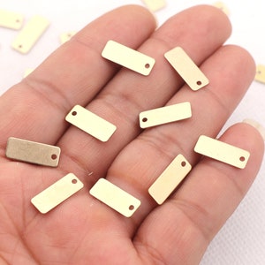 Könnte beinhalten: Eine Sammlung kleiner, goldfarbener Metallanhänger mit einem Loch oben. Die Anhänger sind rechteckig und haben eine glatte, glänzende Oberfläche.