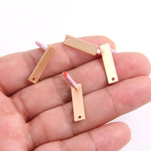 May include: Four gold-colored metal rectangular tags with a hole in each corner. The tags are on a hand.