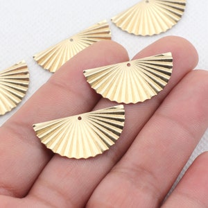 Puede incluir: Encantos de metal dorado en forma de abanico con un agujero en el centro. Los encantos están dispuestos en una mano.