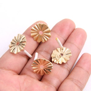 Pode incluir: Quatro costas de brincos de metal dourado com uma borda festonada e um pequeno furo no centro. As costas são mostradas em uma mão.
