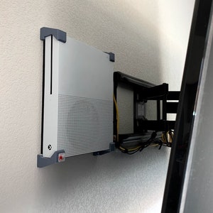 May include: A white gaming console mounted on a wall with gray brackets. The console is connected to a black TV mount with yellow cables.