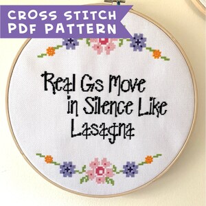May include: A white embroidery hoop with a cross stitch design. The design features a floral border and the text "Real Gs Move in Silence Like Lasagna".