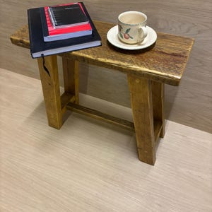 May include: A small, wooden bench with a stack of notebooks and a teacup on a saucer. The bench is made of light brown wood and has a simple design. The teacup has a floral pattern. The bench is approximately 60 cm long.