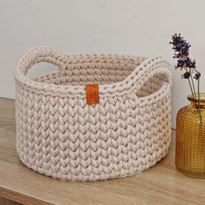 May include: A beige crocheted storage basket with two handles. The basket has a woven texture and a leather label that says "VALTUA".