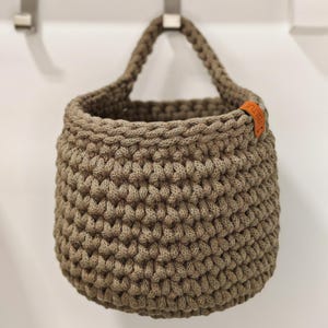 Pu&ograve; includere: Un cesto appeso in crochet marrone con un'etichetta in pelle che dice "Knit" in lettere arancioni.