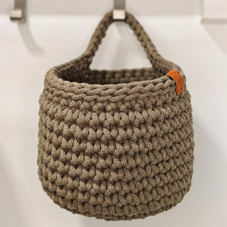 Pu&ograve; includere: Un cesto appeso in crochet marrone con un'etichetta in pelle che dice "Knit" in lettere arancioni.
