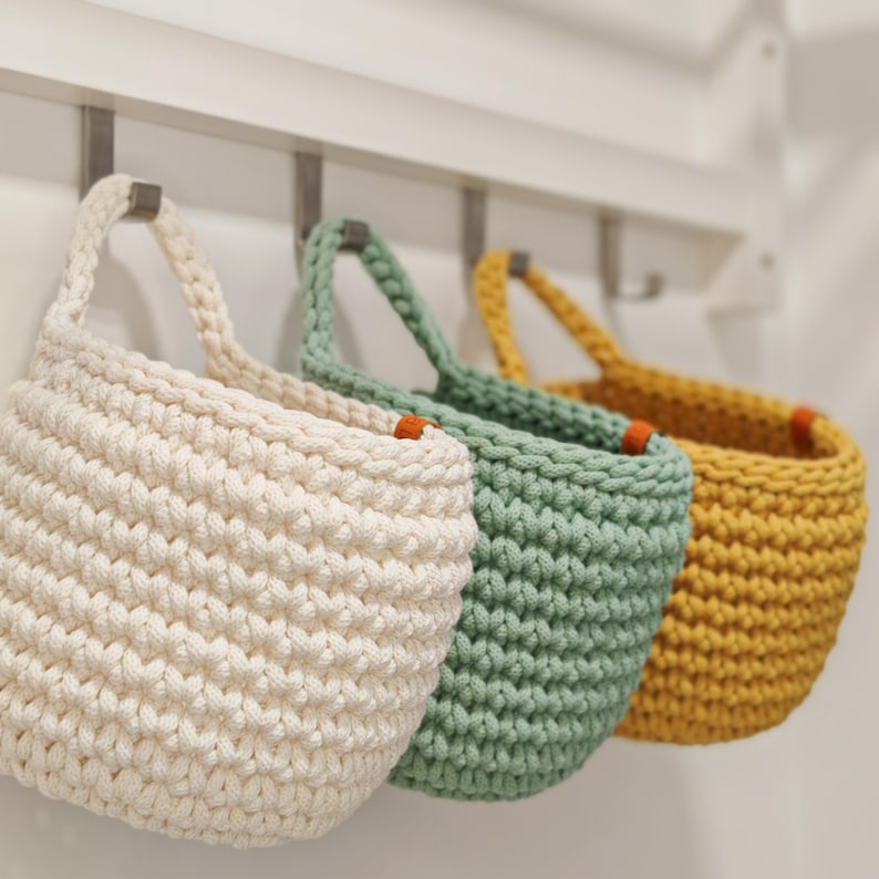 Pu&ograve; includere: Tre cesti appesi all'uncinetto in bianco, verde e giallo. I cesti sono realizzati con filato spesso e hanno un aspetto testurizzato e intrecciato. Sono appesi a ganci su una parete bianca.