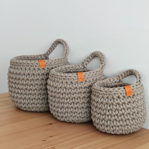 Pu&ograve; includere: Tre cesti appesi in crochet beige con etichette in pelle. I cesti sono di diverse dimensioni e sono appesi a un muro.