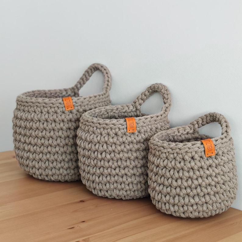 Pu&ograve; includere: Tre cesti appesi in crochet beige con etichette in pelle. I cesti sono di diverse dimensioni e sono appesi a un muro.