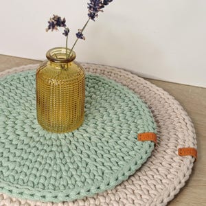 以下が含まれることがあります： 緑とベージュの2つの丸い編み物のコースター。緑のコースターの上に、乾燥したラベンダーの花が入った小さな琥珀色のガラスの花瓶が置かれています。