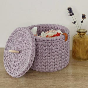 Op de afbeelding: Een lavendelkleurige gehaakte opbergmand met een deksel en een houten handvat. De mand is gevuld met kleine pakjes snoep.