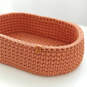 Può includere: Un cestino ovale lavorato a mano all'uncinetto, di colore terracotta caldo. Il cestino presenta un motivo a punto strutturato e un bordo intrecciato. Una piccola etichetta in pelle marrone con la scritta "VALTURIA" è attaccata.