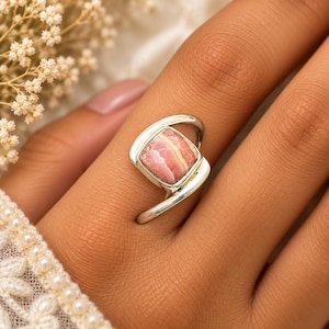 May include: A silver ring featuring a square-shaped, polished rhodochrosite gemstone with distinct pink and white banding. The metal band has a smooth, curved design that wraps partially around the stone, creating an open, asymmetrical setting.