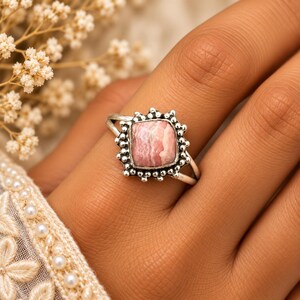 May include: A silver ring featuring a square-cut, pink and white banded rhodochrosite gemstone. The stone is set in a decorative silver bezel surrounded by a border of small, raised silver spheres, mounted on a simple, polished silver band.